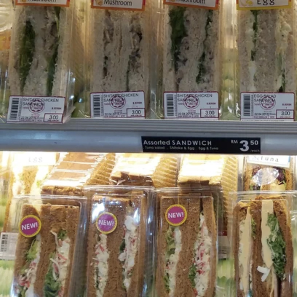 Packaged sandwiches are displayed on refrigerated shelves, labeled "Shitake Chicken," "Mushroom," and "Egg." The bottom shelf has a sign reading "Assorted SANDWICH RM 3.50 each," with some packs labeled "NEW!"