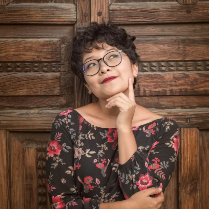 Person wearing glasses and a floral dress, thoughtfully resting their chin on their hand, standing against a rustic wooden backdrop with a decorative pattern.