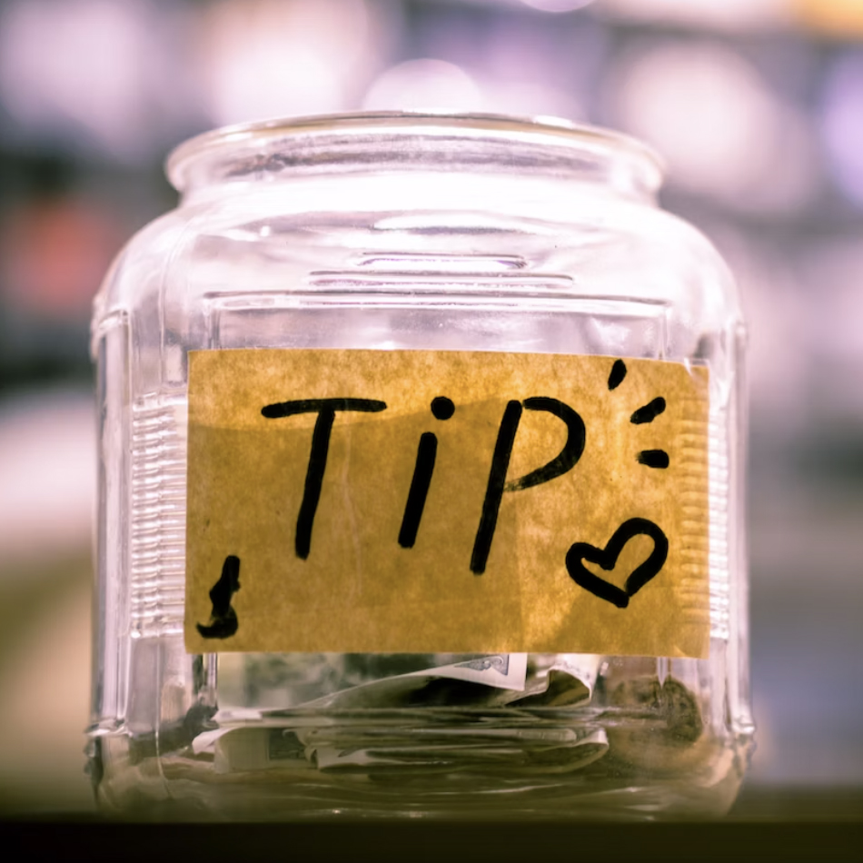 A clear jar labeled "TIP" with money inside sits on a counter in a blurred background, suggesting a cafe or store environment.