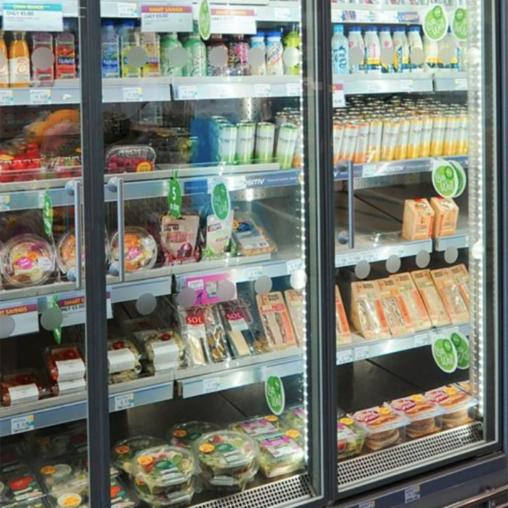 A refrigerated supermarket display case contains prepared salads, sandwiches, drinks, and packaged meals, all organized on shelves with price tags and green promotional signage visible.