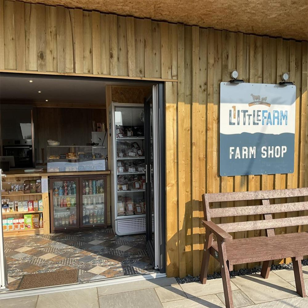 Store counter displays various goods; behind glass are baked items, below are beverages. The setting is a wooden farm shop with a sign reading “LITTLE FARM FARM SHOP” near a bench outside.