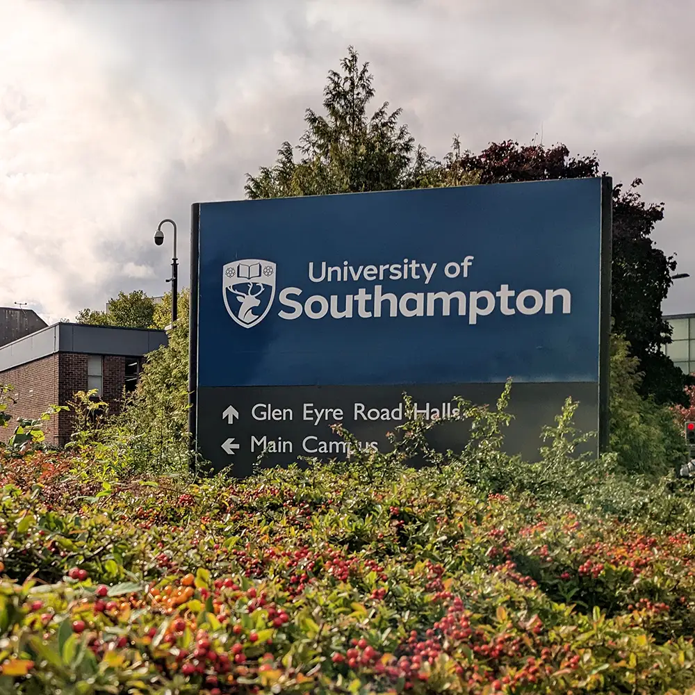 A blue sign reads "University of Southampton" with directional arrows pointing to Glen Eyre Road Halls and Main Campus, surrounded by green shrubs, against a backdrop of buildings and trees.