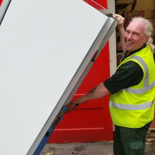 A man in a yellow safety vest lifts a large, white, rectangular object in front of a bright red door, smiling as he works. He appears to be outside or in an open garage.