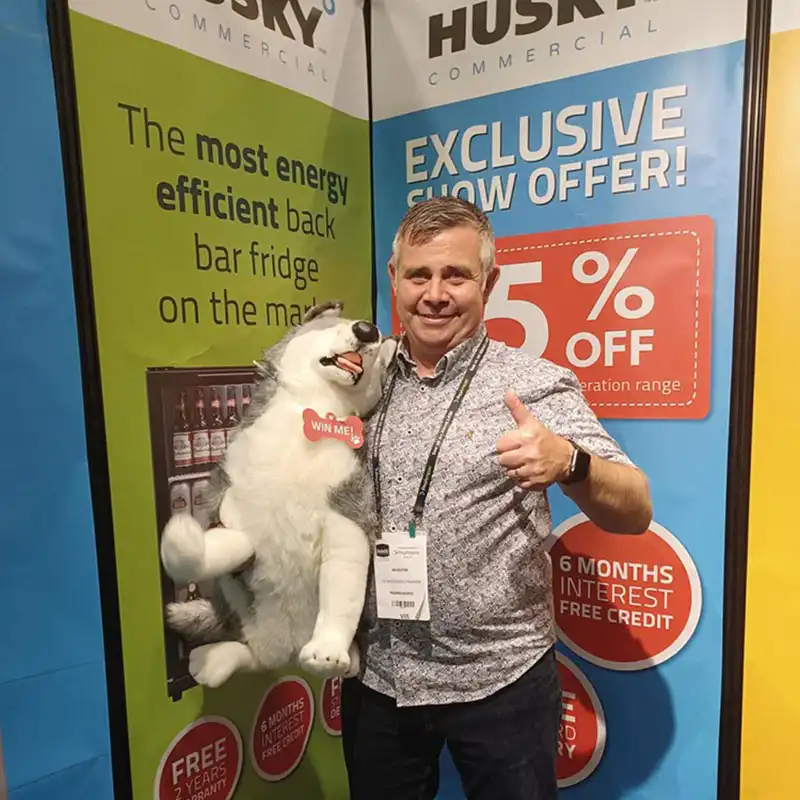 A smiling man, holding a plush husky toy, gives a thumbs up in front of colorful promotional signage displaying "WIN ME!" on the toy and "25% OFF" alongside details of interest-free credit.