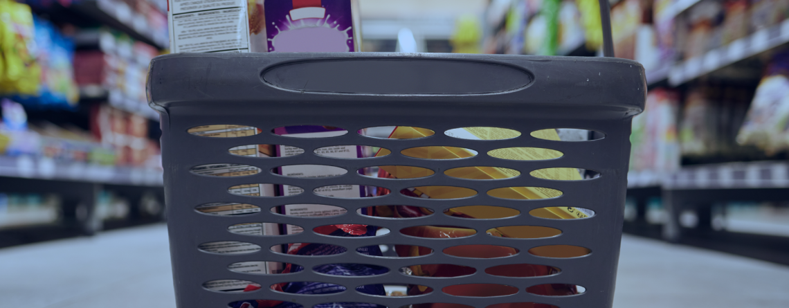 Grocery shopping basket in a supermarket aisle with shelves and a commercial display fridge in the background