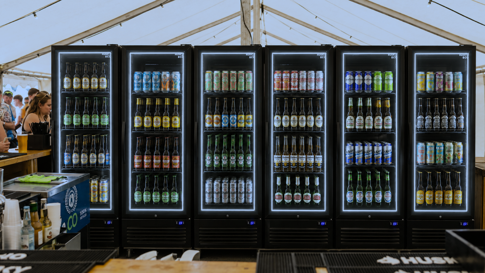 Husky drinks fridges stocked with beers and canned beverages at a busy festival bar, showcasing illuminated commercial drinks fridges for high-volume event service.